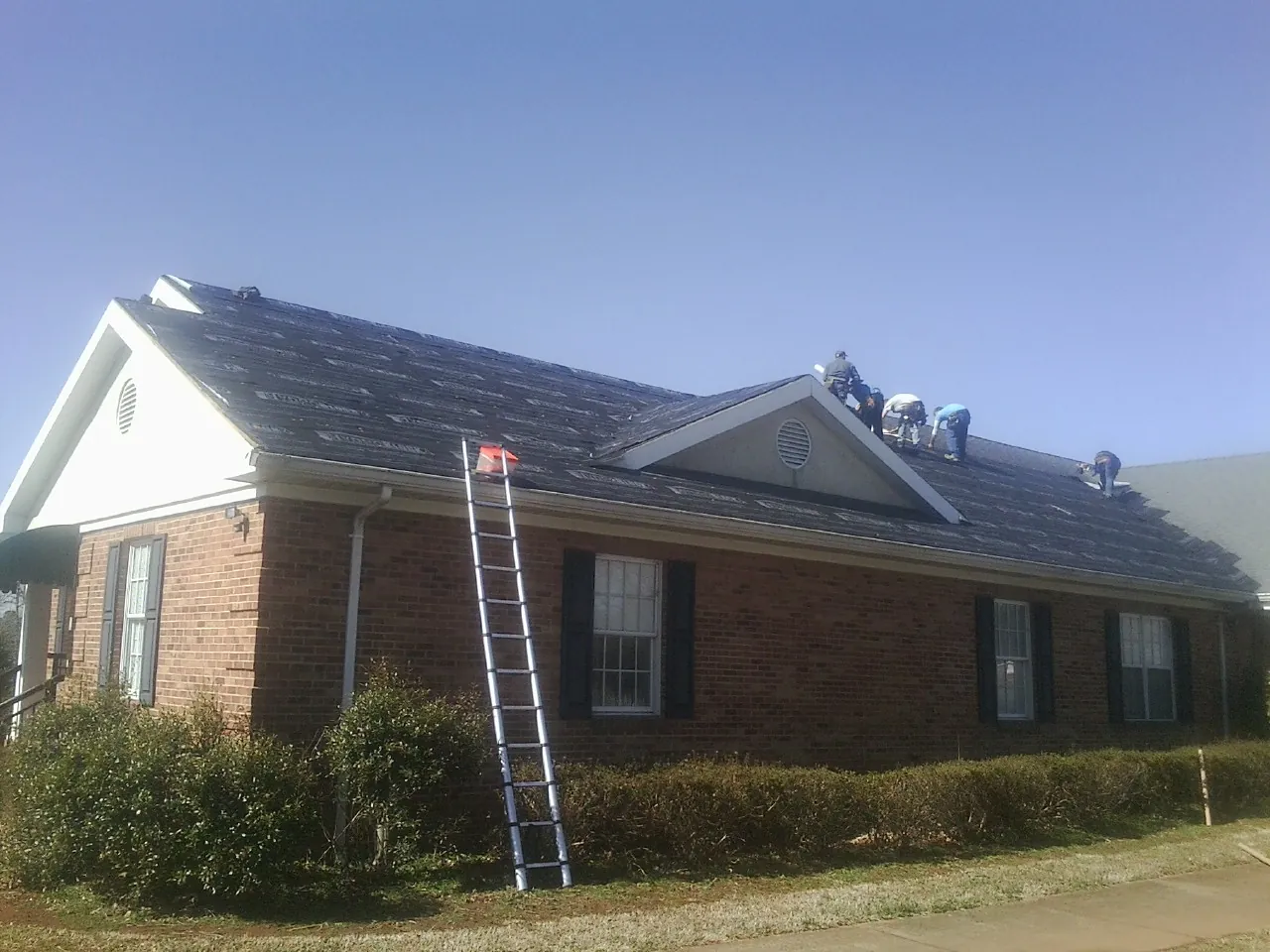 Crew tearing shingles off a brick ranch roof with a ladder set — Emergency Roof Repair work by our crew in Bayard