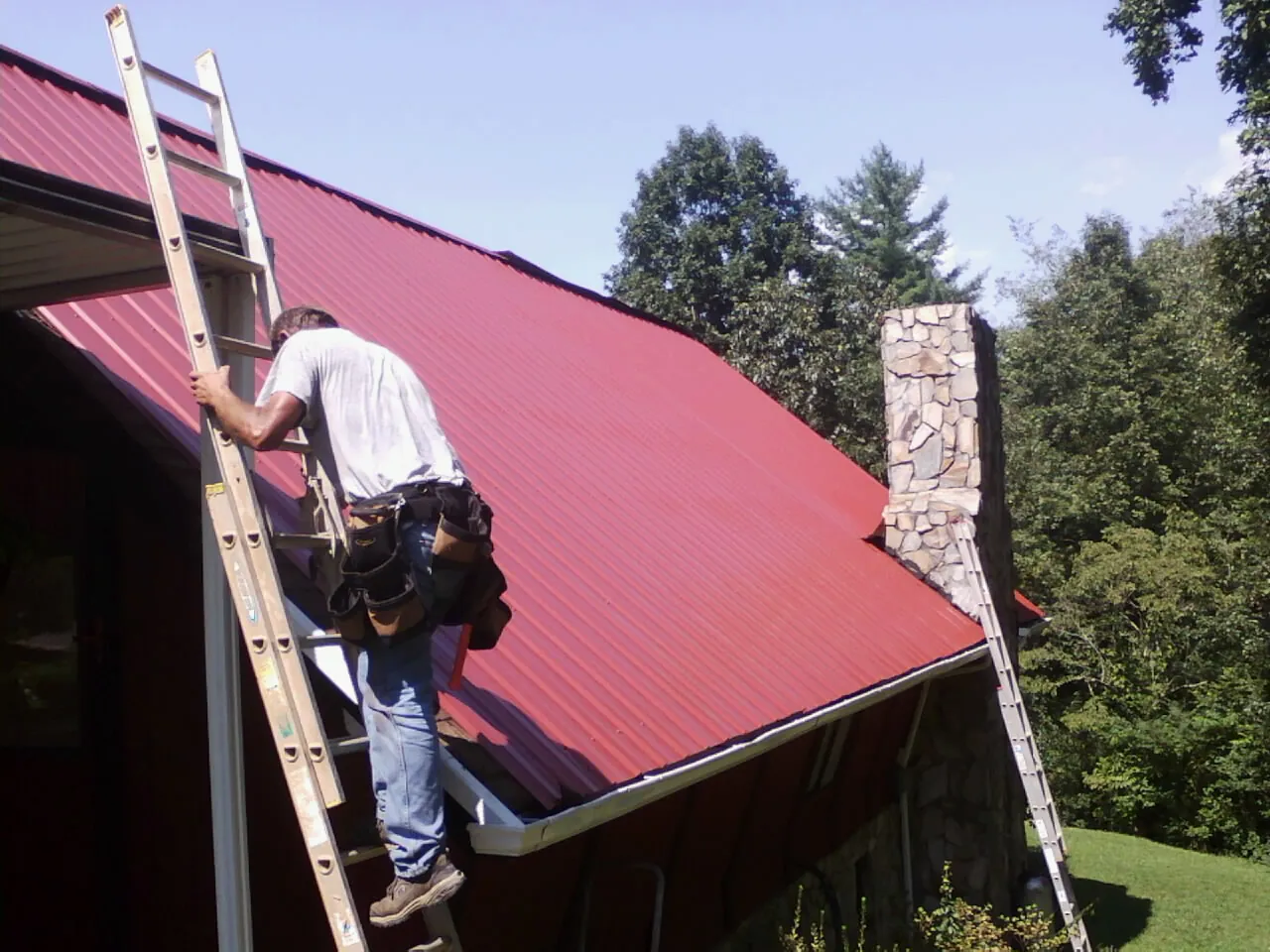 Roofer on a ladder beside a red metal roof and stone chimney — Soffit & Fascia Repair by Roofing Brothers in Bayard